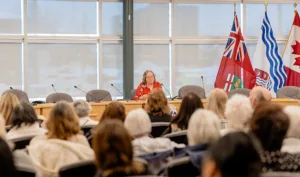 Woman speaking in front of a seated crowd in conference room.